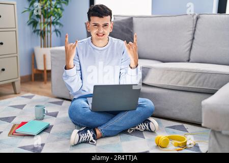 Nicht binäre Person, die mit einem Computer-Laptop lernt, auf dem Boden sitzt und mit verrücktem Ausdruck schreit und mit erhobenen Händen ein Rocksymbol macht. Musikstar. in Stockfoto