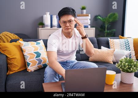 Ein junger asiatischer Mann, der zu Hause ein Notebook benutzt, auf dem Sofa sitzt und sich erschossen und Hand und Finger an den Kopf zeigt, wie eine Waffe, Selbstmordgeste. Stockfoto