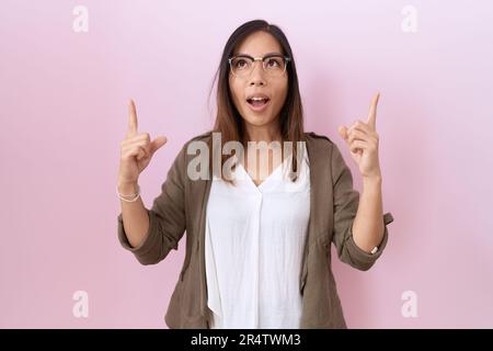 Eine chinesin mittleren Alters, die eine Brille auf pinkfarbenem Hintergrund trug, war erstaunt und überrascht, als sie aufblickte und mit Fingern und erhobenen Armen zeigte. Stockfoto