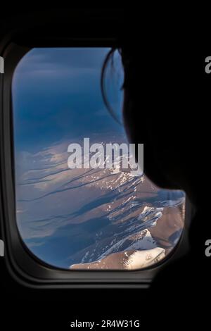 Silhouette einer Person, die von ihrem Sitz aus aus aus aus einem Flugzeugfenster schaut. Kaukasusgebirge - Luftaufnahme bei Nacht aus einem Flugzeug Stockfoto