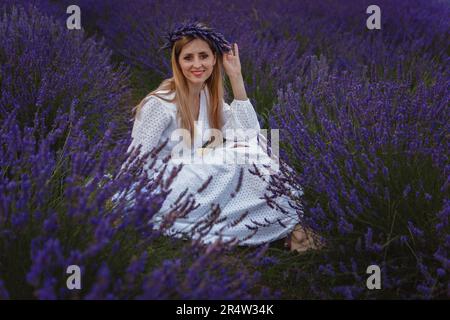 Eine junge Frau in einem weißen Kleid sitzt in der Mitte eines Lavendelfeldes Stockfoto