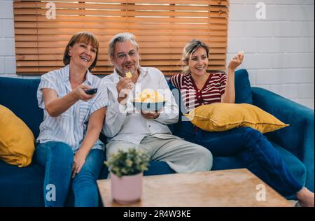 Familienmitglieder aus mehreren Generationen genießen eine schöne Zeit zusammen, indem sie Fernsehen, Snacks und Gespräche teilen und neue entdecken Stockfoto