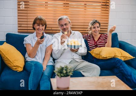 Familienmitglieder aus mehreren Generationen genießen eine schöne Zeit zusammen, indem sie Fernsehen, Snacks und Gespräche teilen und neue entdecken Stockfoto