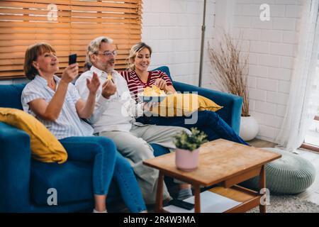 Familienmitglieder aus mehreren Generationen genießen eine schöne Zeit zusammen, indem sie Fernsehen, Snacks und Gespräche teilen und neue entdecken Stockfoto