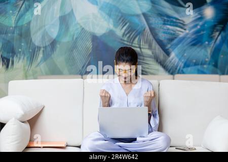 Überglücklich, junge Frau, die auf der Couch sitzt und den Erfolg feiert Stockfoto