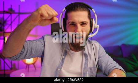 Ein bärtiger Gamer in einem Gaming-Headset gewinnt ein Videospiel. Cybersportler in einem Neonraum. Lifestyle-Gamer, Online-Spiele. Sieg im E-Sport Stockfoto