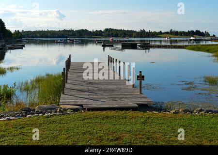 Ein langer hölzerner Bootssteg, der in einen Teich ragt. Es gibt mehrere Boote, die im Wasser vertäut sind. Es gibt großes grünes Gras an der Küste. Stockfoto