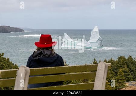 Eine Frau sitzt auf einer Bank und trägt einen lebendigen roten Hut und eine schwarze Jacke mit Blick auf einen großen weißen Gletschereisberg, der im kaltblauen Ozean schwimmt. Stockfoto