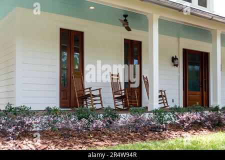 Drei altbraune Schaukelstühle aus Holz auf der Veranda eines alten weißen Landhauses mit einer umlaufenden Veranda. Da ist ein Blumenbeet. Stockfoto