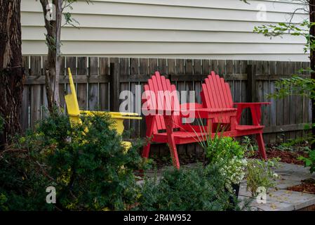 Vintage cremefarbene Cape Cod horizontale Wand und ein Holzzaun. Es gibt einen gelben Adirondack-Stuhl und zwei rote zusammengesetzte Gartenstühle. Stockfoto