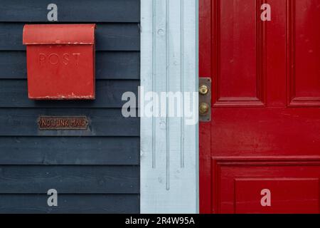 Ein hellroter Briefkasten oder Briefkasten aus Metall an einer dunkelblauen Holzwand mit einer roten Tür. Die Mailbox hat eine gebogene Abdeckung. Da ist ein glänzender Türgriff Stockfoto