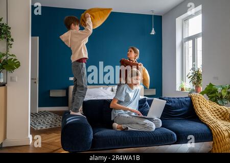 Mutter, die an einem Laptop arbeitet, während ihre aktiven, lauten Kinder mit Kissen spielen und Spaß haben Stockfoto