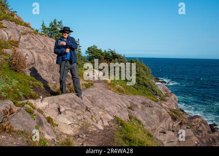 Senior männlich wandern entlang eines Pfades am Rande eines Berges. Der Pfad liegt am Rand einer hohen Klippe mit Blick auf den blauen Ozean und den Himmel. Der Mann Stockfoto