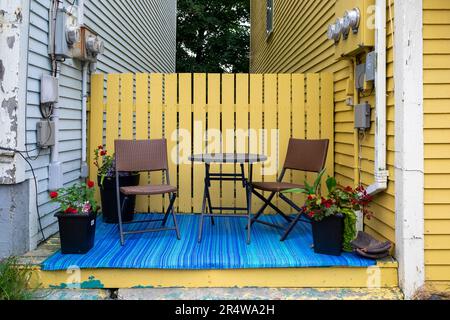 Zwei kleine Terrassenstühle und ein runder Tisch zwischen zwei Häusern mit gelbem Zaun, Blumen und blauer Matte. Stockfoto