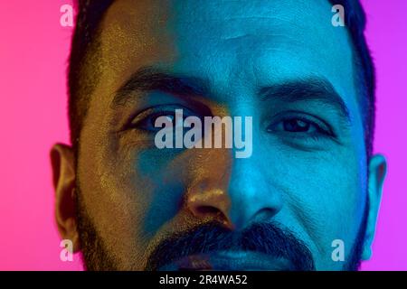 Nahaufnahme des zugeschnittenen männlichen Auges, das mit Ruhe und Aufmerksamkeit vor dem Farbverlauf eines rosa, blauen Studiohintergrunds in Neonlicht schaut Stockfoto
