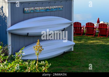 Zwei weiße Kajaks hängen an einem kleinen grauen Bootsschuppen. Da ist ein Schild über dem Gebäude. Drei rote Stühle stehen auf Gras und blicken zum Meer. Stockfoto