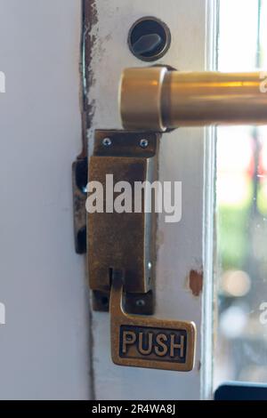 Ein Schiebebügelöffner aus altem Messing auf einer weißen Holztür mit einem großen Glasfenster. Der verschlissene Griff hat das Wort „Drücken“ auf einem verschlissenen Metallknopf. Stockfoto