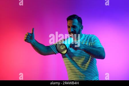Ein bärtiger glücklicher Mann, der in Megafon spricht und eine positive Geste zeigt, mit den Händen gegen den Farbverlauf, den rosafarbenen, blauen Studiohintergrund in Neonlicht Stockfoto