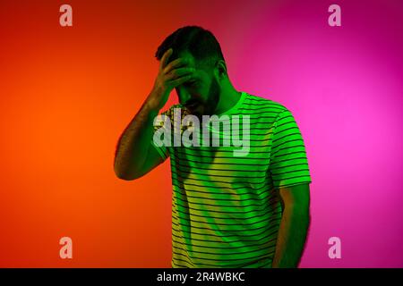 Porträt eines bärtigen Mannes, der die Hand auf der Stirn hält und mit einem durchdachten Gesicht vor dem Hintergrund des Gradient Studio in Neonlicht posiert Stockfoto