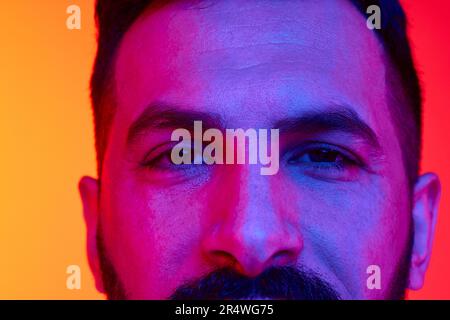 Nahaufnahme des zugeschnittenen männlichen Auges, das mit Ruhe und Aufmerksamkeit vor einem verlaufenen roten, orangefarbenen Studiohintergrund in Neonlicht schaut Stockfoto