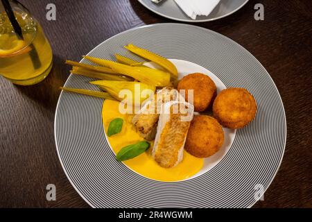 Filet vom Kabeljau, Polenta-Krocket in Karotten- und Orangensaft, marinierter Fenchel auf gestreiftem Teller. Limonade und ein Teil des zweiten Tellers im Restaurant t Stockfoto