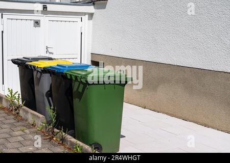 Mehrfarbige Kunststoffbehälter zur separaten Abfallsammlung, die in einer Reihe neben der Garage stehen. Stockfoto