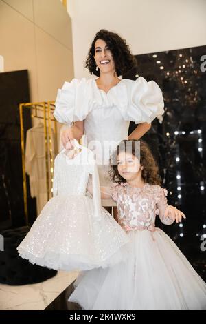 Charmante Braut aus dem Nahen Osten mit braunem Haar, die in einem weißen Hochzeitskleid mit Puffärmeln und Rüschen steht und ein Mädchenkleid mit Tullerock hält Stockfoto