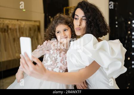 Nahöstliche Braut mit braunem Haar in weißem Hochzeitskleid mit Puffärmeln und Rüschen, die Selfie auf dem Smartphone macht, während sie fast glücklich Lippen schmollt Stockfoto