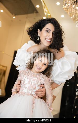Charmante Braut aus dem Nahen Osten mit braunem Haar in weißem Hochzeitskleid, Puffärmel und Rüschen hinter der süßen Tochter und lächelnden Beiden Stockfoto