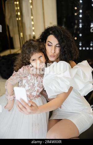 Lustige Braut aus dem Nahen Osten mit braunem Haar in weißem Hochzeitskleid mit Puffärmeln und Rüschen, die Selfie mit ihrer Tochter auf dem Smartphone macht, während sie unterwegs ist Stockfoto