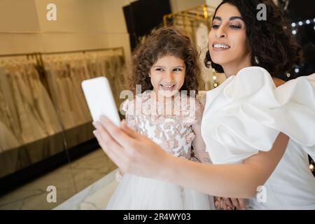 Charmante Braut aus dem Nahen Osten mit braunem Haar in weißem Hochzeitskleid mit Puffärmeln und Rüschen, die Selfie mit ihrer glücklichen Tochter auf dem Smartphone macht Stockfoto