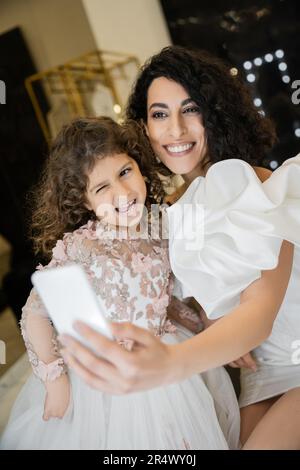 Charmante Braut aus dem Nahen Osten mit braunem Haar in weißem Hochzeitskleid mit Puffärmeln und Rüschen, die Selfie auf dem Smartphone mit einer lustigen Tochter macht Stockfoto