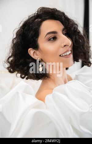 Portrait einer fröhlichen braunen Braut aus dem Nahen Osten mit welligen Haaren, die in trendigen Hochzeitskleid mit Puffärmeln und Rüschen in Brautboutique und Klo posieren Stockfoto