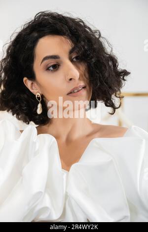Porträt einer braunen Braut aus dem Nahen Osten mit welligen Haaren, die in trendigen Hochzeitskleidern posieren, mit Puffärmeln und Rüschen in einer Brautboutique und einem Look Stockfoto