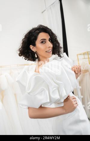 Glückliche Braut aus dem Nahen Osten mit braunem und welligem Haar, die mit Händen auf der Hüfte posiert, in einem trendigen Hochzeitskleid mit Puffärmeln und Rüschen in Brautbuti Stockfoto