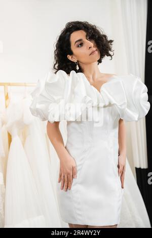 Elegante Braut aus dem Mittleren Osten mit braunem und welligem Haar in trendigen Hochzeitskleid mit Puffärmeln und Rüschen in Brautboutique neben Tulle Stockfoto