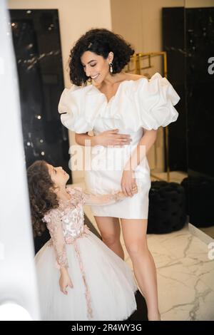 Ein fröhliches und süßes kleines Mädchen aus dem Nahen Osten in Blumenkleidung, das die Braut mit welligen Haaren ansieht und in einem trendigen Hochzeitskleid mit Puffärmel und Rüschen steht Stockfoto