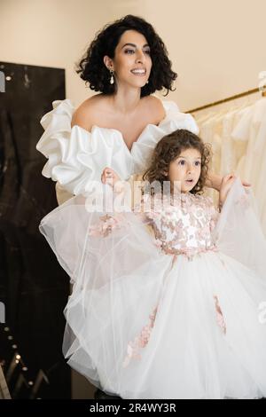 Faszinierende Braut aus dem Nahen Osten mit braunem Haar, die in einem weißen Hochzeitskleid mit Puffärmel und Rüschen steht und wegschaut, während sie Tulle hält Stockfoto