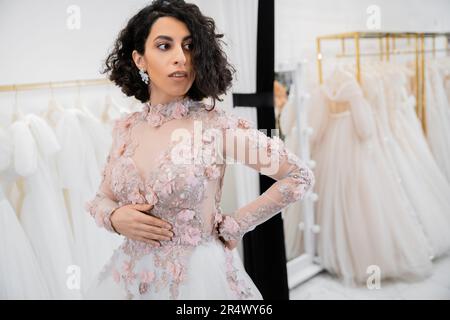 Braut, verführerischer naher Osten und brünette Frau mit welligen Haaren in einem wunderschönen und blumigen Hochzeitskleid in einem luxuriösen Salon Stockfoto