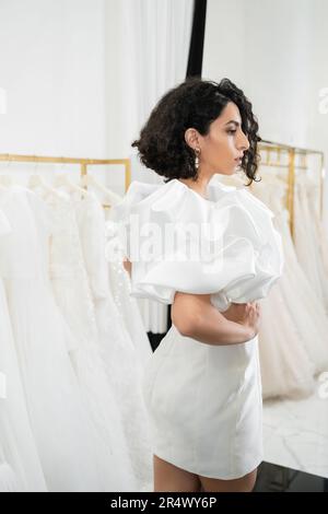 Braune Braut aus dem Nahen Osten mit braunem und welligem Haar in trendigen Hochzeitskleid mit Puffärmeln und Rüschen in der Brautboutique neben tull Stockfoto