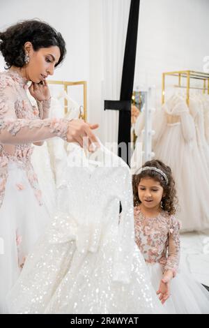 Brünette Braut aus dem Nahen Osten in geblümtem Hochzeitskleid, die ihr hilft, das Kleid für ihre kleine Tochter im Brautsalon zu wählen, rund um weiße Tulle-Stoffe, Proce Stockfoto
