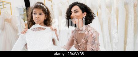 Eine bezaubernde und brünette Braut aus dem Nahen Osten in einem blumigen Hochzeitskleid hilft ihr dabei, das Kleid für ihre kleine Tochter im Brautsalon in weißer Tüle auszuwählen Stockfoto