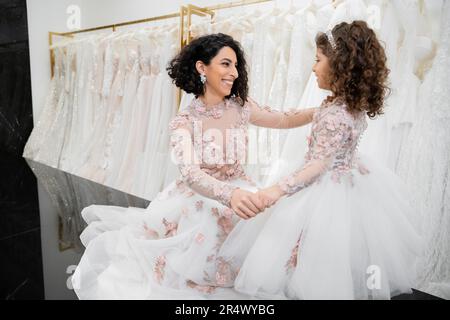 Besonderer Moment, glückliche Braut aus dem Nahen Osten im Hochzeitskleid, die mit ihrer kleinen Tochter im Brautsalon um weiße Tulle Fabri sitzt und Händchen hält Stockfoto