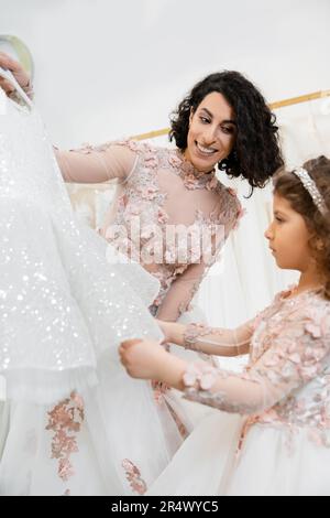 Charmante und brünette Frau aus dem Nahen Osten in geblümtem Hochzeitskleid, die ihr bei der Auswahl des Kleides für ihre kleine Tochter im Brautsalon um weiße Tulle hilft Stockfoto
