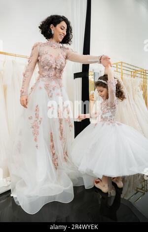 Eine glückliche Frau aus dem Nahen Osten in einem blumigen Hochzeitskleid, die mit einem lächelnden Mädchen in süßer Kleidung tanzt, mit einem Tulle-Rock im Brautsalon, Shopping und einem besonderen Moment Stockfoto