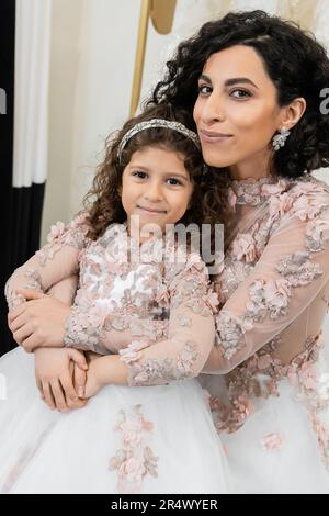 Fröhliche Braut aus dem Nahen Osten im Hochzeitskleid, die ein fröhliches Mädchen in süßer Blumenkleidung umarmt, im Brautsalon, beim Einkaufen, bei einem besonderen Moment, Mutter und Tochter Stockfoto