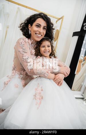 Eine reizende Braut aus dem Nahen Osten im Hochzeitskleid, die ein glückliches Mädchen in süßer Blumenkleidung umarmt, im Brautsalon, beim Einkaufen, bei einem besonderen Moment, bei Mutter und Tochter Stockfoto