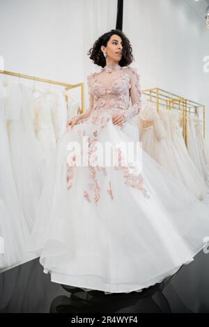 Eine brünette Frau aus dem Nahen Osten mit welligen Haaren, die ein wunderschönes und geblümtes Hochzeitskleid anprobiert, in einem luxuriösen Brautsalon, Einkaufen Stockfoto
