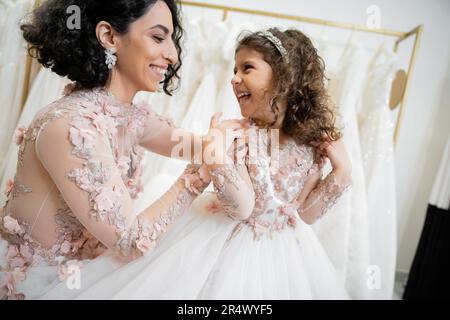 Glückliche Frau aus dem Nahen Osten mit braunem Haar in geblümtem Hochzeitskleid, die die Schultern einer lächelnden Tochter in süßer Kleidung umarmt, mit Tullerock in der Braut Stockfoto