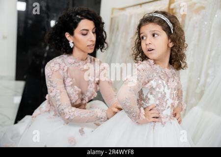 Verschwommene Braut aus dem Nahen Osten mit braunem, welligem Haar im Hochzeitskleid, passend für das süße Blumenkleid einer überraschten Tochter im Brautsalon, beim Einkaufen, bei Spe Stockfoto